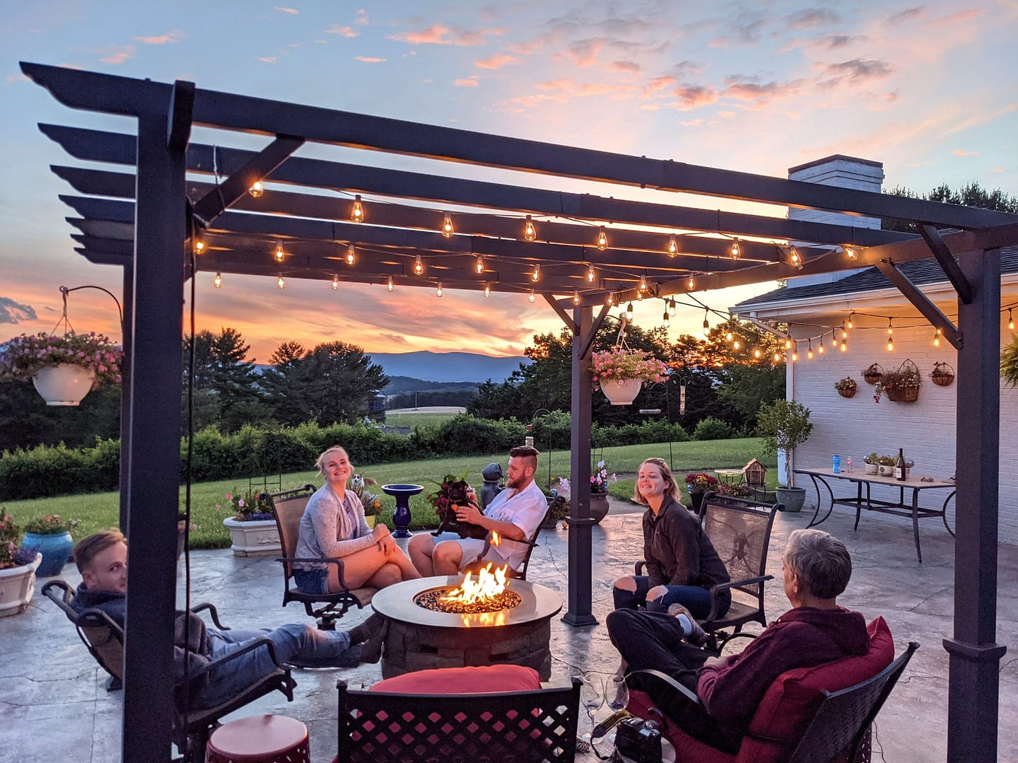 A family relaxing around a fire table in outdoor seating area under pergola with lights at sunset