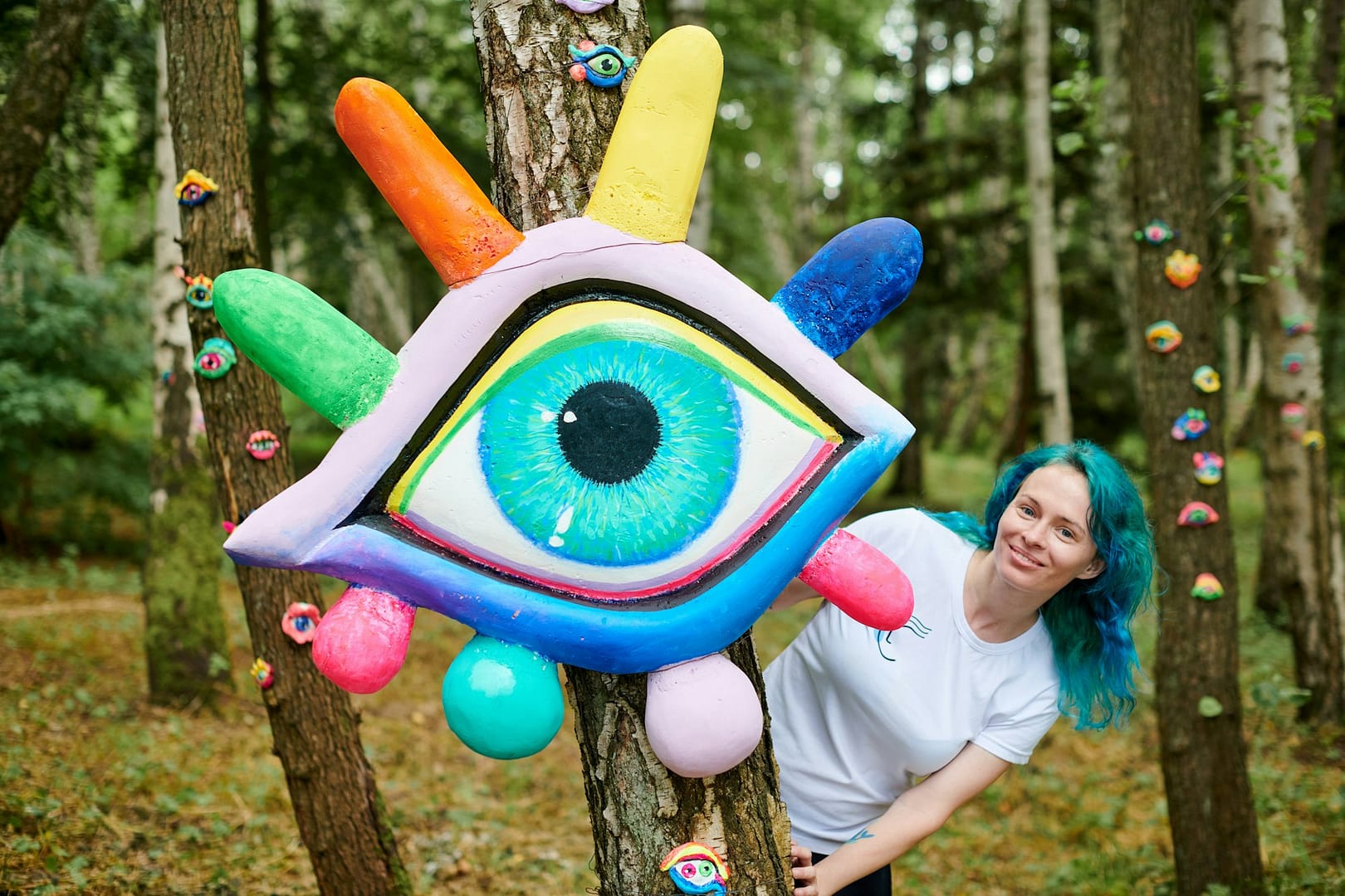 Smiling young adult woman with dyed turquoise hair in white T shirt near big eye art installation